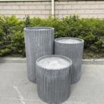 A collection of Fluted Garden Pots in raw concrete color, showcasing their elegant ribbed design. The pots are available in various sizes, including small, medium, and large, and are arranged in a visually appealing cluster. The textured surface and sleek finish of the pots add a touch of sophistication to the garden setting.